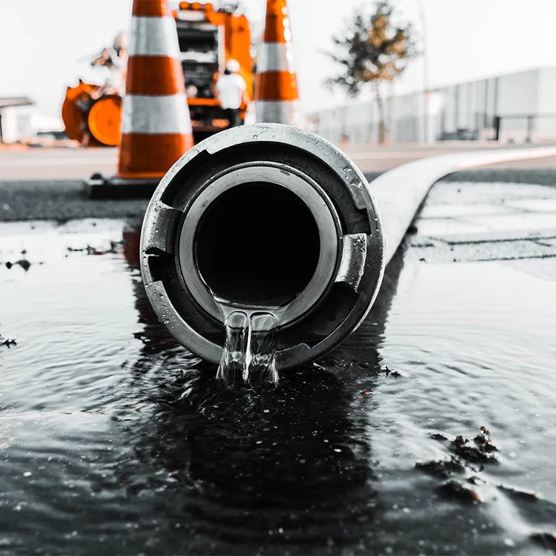 Close-up view of water flowing from a large pipe on a flooded street, with emergency orange traffic cones and repair equipment in the background. Alton Facility Services, serving Hampshire, Surrey, and the UK