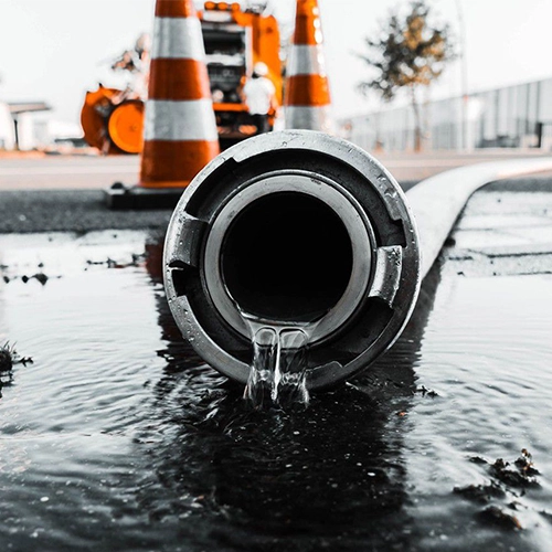 Close-up view of water flowing from a large pipe on a flooded street, with emergency orange traffic cones and repair equipment in the background. Alton Facility Services, serving Hampshire, Surrey, and the UK